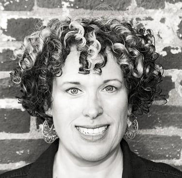 Heather Blaikie, a woman with curly hair and a black jacket standing in front of a brick wall