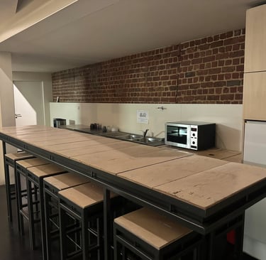 Cuisine de coworking avec longue table haute en bois, tabourets, évier, micro-ondes et mur en briques apparentes.