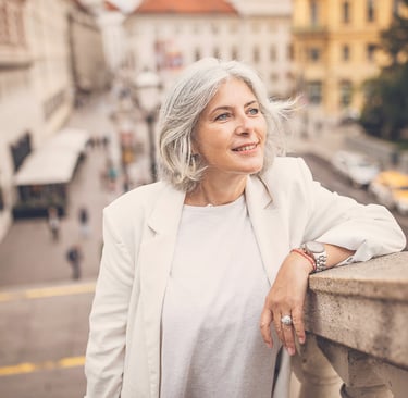 Smiling senior woman with gray hair wearing a white blazer while traveling in a European city.