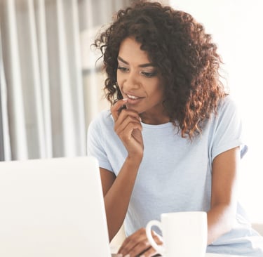 A smiling African American woman with curly hair working on a laptop at home with a coffee mug.