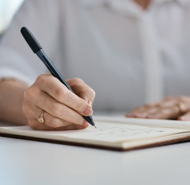 Close-up of a woman’s hand writing in an open journal with a black pen, capturing a quiet moment.