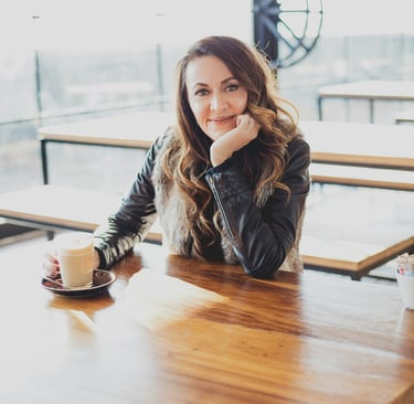 Smiling woman in a black leather jacket enjoying a latte coffee at a bright modern cafe table.