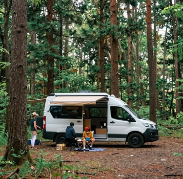 A rustic wooden mobile home is parked at the side of a quiet street with minimal traffic. The home has a triangular roof adorned with colorful pennant flags, and there are small windows on the back. The street is lined with traditional buildings and a stone wall, and the sky is partly cloudy, indicating a sunny day.