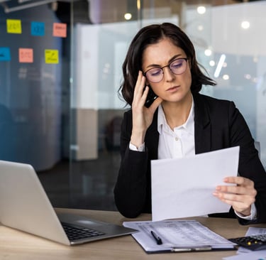 Stressed businesswoman in glasses reviewing financial documents at her office desk with a laptop.