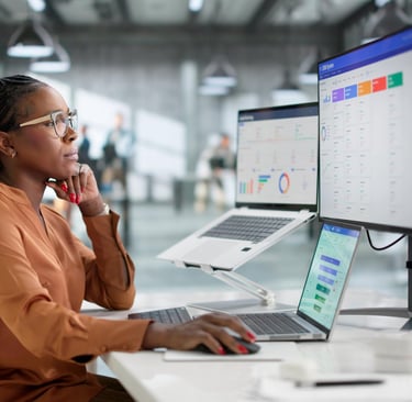 A female data analyst reviews a CRM system dashboard on multiple monitors in a modern office.