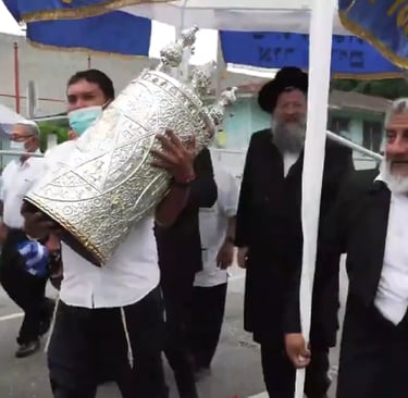 Jewish men carry a silver Torah scroll during a festive outdoor parade under a blue and white canopy.