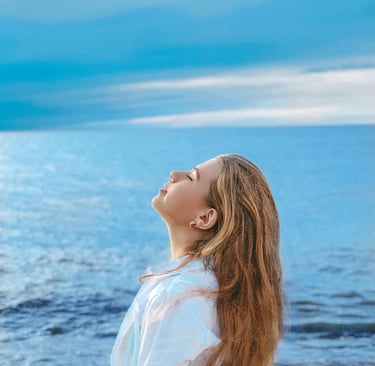 A young woman with long hair enjoys a peaceful moment by the blue ocean waves under a cloudy sky.