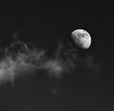 A black-and-white photograph of a line of cloud leading up to a gibbous moon.