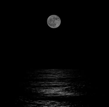 A black-and-white photograph of the full moon rising above the ocean and its moonlight on the water in Cape May, NJ.