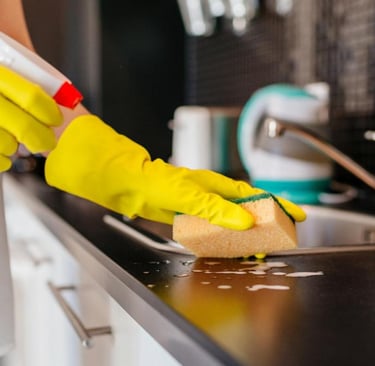 A person wearing yellow rubber gloves uses a sponge and spray cleaner to sanitize a kitchen countertop.