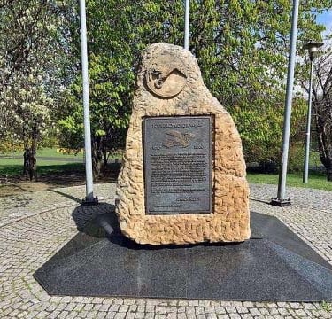 The obelisk for Mokotow Airport, in Warsaw, Poland. Sandy yellow coloured shaped rock with metal plaque inserted, in a park.