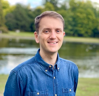 Chris Philpot, a white man in his 30s, smiling at the camera. He is wearing a blue denim shirt and standing in a park.