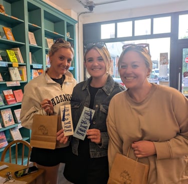 Three smiling women holding shopping bags and linocut bookmarks inside a cozy local bookstore.