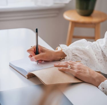 Ein aufgeschlagenes Notizbuch auf einem Tisch. Eine Frau schreibt mit dem Stift etwas in das Buch.