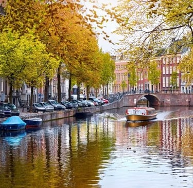 Tour boat cruising on an Amsterdam canal lined with historic buildings and autumn trees.