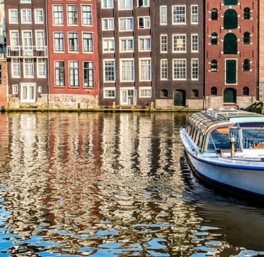 A boat tours the Amsterdam canal beside colorful historic Dutch townhouses reflecting in the water.