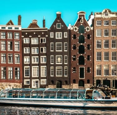 A glass-roofed canal boat cruises past iconic historic gabled houses in Amsterdam under a clear blue sky.