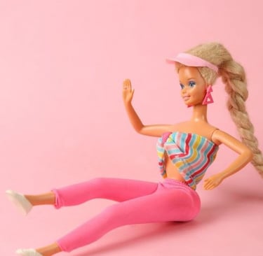 Blonde Barbie doll in 80s workout clothes with pink leggings and striped top against a pink background.