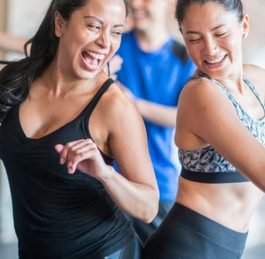 Two smiling women in activewear enjoying a high-energy dance fitness class at a gym.