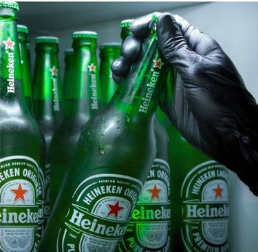 A gloved hand grabbing a cold bottle of Heineken lager beer from a refrigerator shelf.