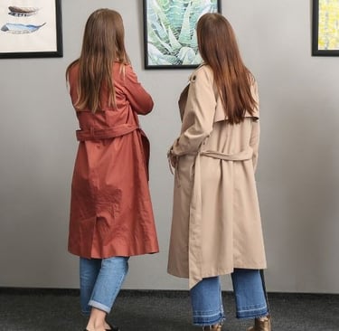 Two women wearing long trench coats in rust orange and beige tan colors while viewing art in a gallery.