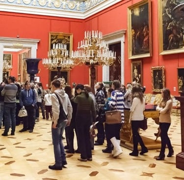 Tourists viewing classic oil paintings in an ornate red art gallery with chandeliers.
