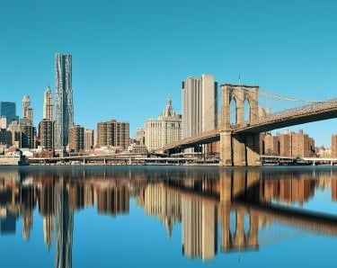 Panoramic Lower Manhattan skyline and Brooklyn Bridge reflecting in the East River under a clear blue sky.