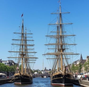 Two historic tall ships with wooden masts docked in a city canal under a clear blue sky.