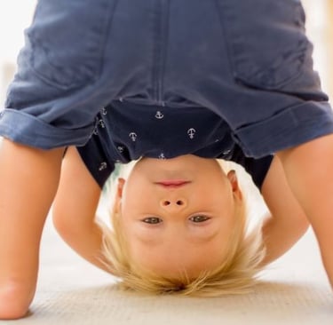 A playful toddler boy with blonde hair looking at the camera while leaning upside down.