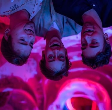 Three smiling men under vibrant pink and purple neon lights. at the upside down museum