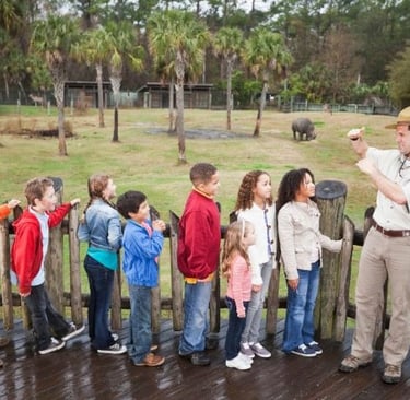 Park ranger teaching a group of children about a rhinoceros at an outdoor zoo exhibit.