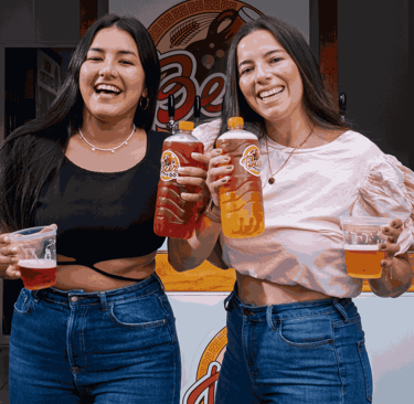 2 mujeres sonriendo sosteniendo vasos y botellas de cerveza artesanal