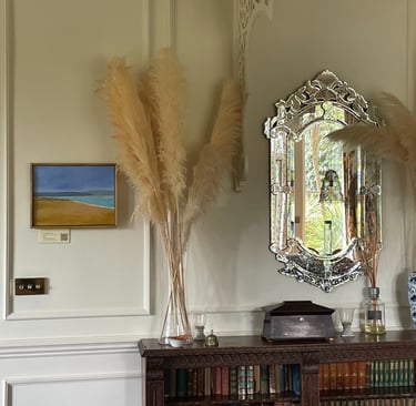 Elegant home decor featuring pampas grass in a glass vase, an ornate Venetian mirror, and a vintage bookshelf.