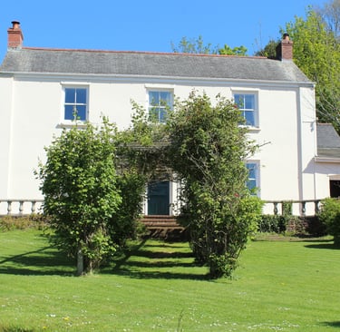 A white two-story country house with a slate roof and green garden hedges.