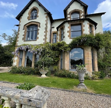 A historic country house featuring gothic windows, stone architecture, and purple wisteria climbing the walls.