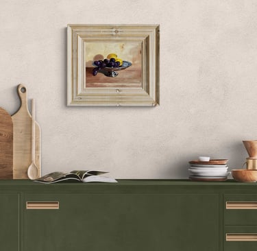 Framed still life oil painting of grapes and lemons hanging above a modern olive green kitchen cabinet.