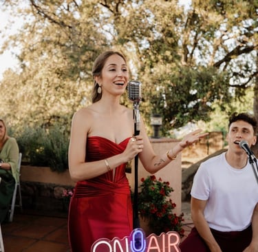 A female singer in a red dress performing with a vintage microphone at an outdoor garden wedding event.