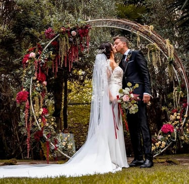 Bride and groom sharing kiss in front of circular wedding arch decorated with red and white flowers during outdoor ceremony.