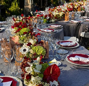 An elegantly set long outdoor dining table with a vibrant floral runner featuring red and white blossoms.