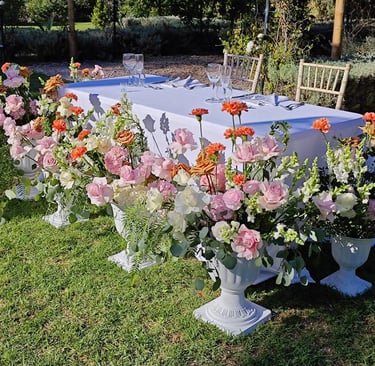 An outdoor wedding table elegantly decorated with lush pink and orange floral arrangements in white urns on a grassy lawn.