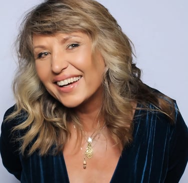 Smiling woman with wavy blonde hair wearing a blue velvet dress and gold necklaces.