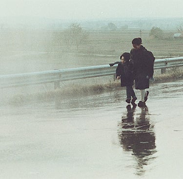 A wide, misty shot of two people walking along a wet, reflective roadside under a hazy grey sky.