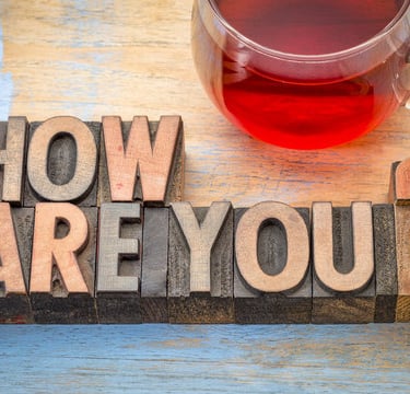 Wooden letterpress blocks spelling How Are You? next to a glass mug of red herbal tea on a rustic table.