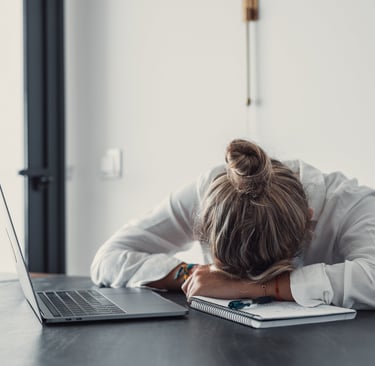 An exhausted woman napping on a notebook next to a laptop, illustrating burnout and work fatigue.