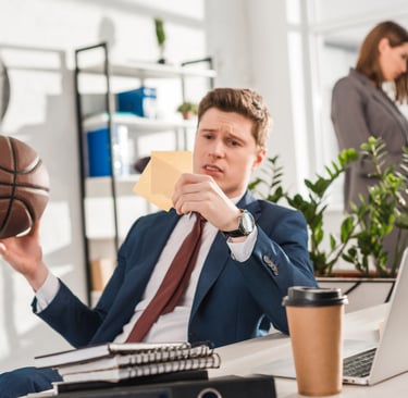 Confused businessman holding a basketball and sticky notes at his office desk with a laptop.