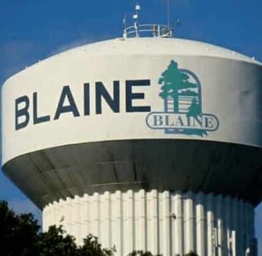 A large white municipal water tower in Blaine, Minnesota, featuring the city logo against a clear blue sky.