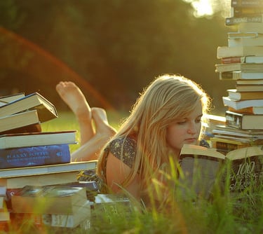 Image of a blonde girl surrounded by books reading a book in the middle of an open field