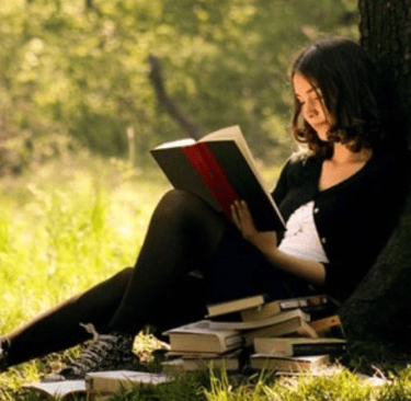 A woman sitting on the ground at the foot of a tree reading a book