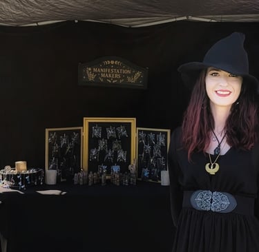 Woman in a black dress and witch hat in front of a Manifestation Makers sign and her jewelry display