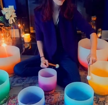 A person seated on a colorful rug is playing crystal singing bowls of different colors and candles 